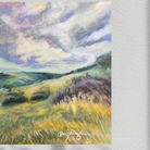 An impressionistic landscape painting of rolling green and yellow hills under a dramatic cloudy sky, displayed on a textured white wall. The artist's signature 'Gretchen Tomasi' is visible in white script at the bottom.