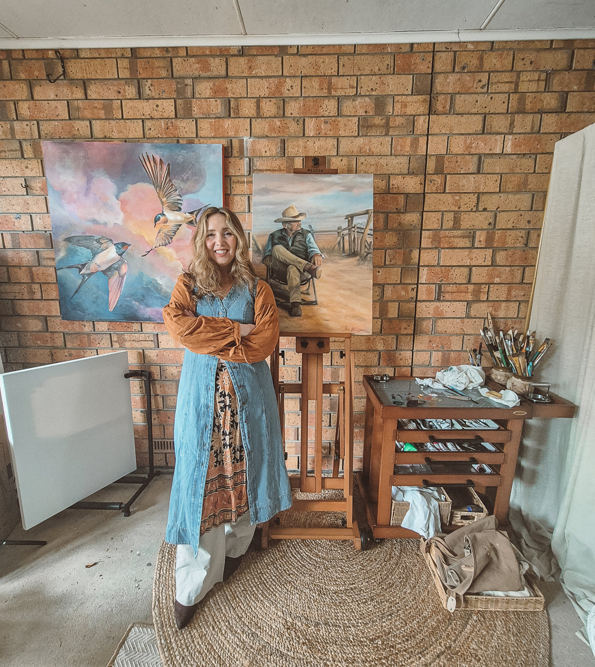 A woman stands in an artist studio wearing a long-sleeved blue dress over an orange top and white pants, posing in front of two paintings on a brick wall.